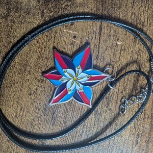 May include: A black cord necklace with a silver metal pendant featuring a colorful star-shaped flower design. The flower has red, blue, white, and yellow accents.