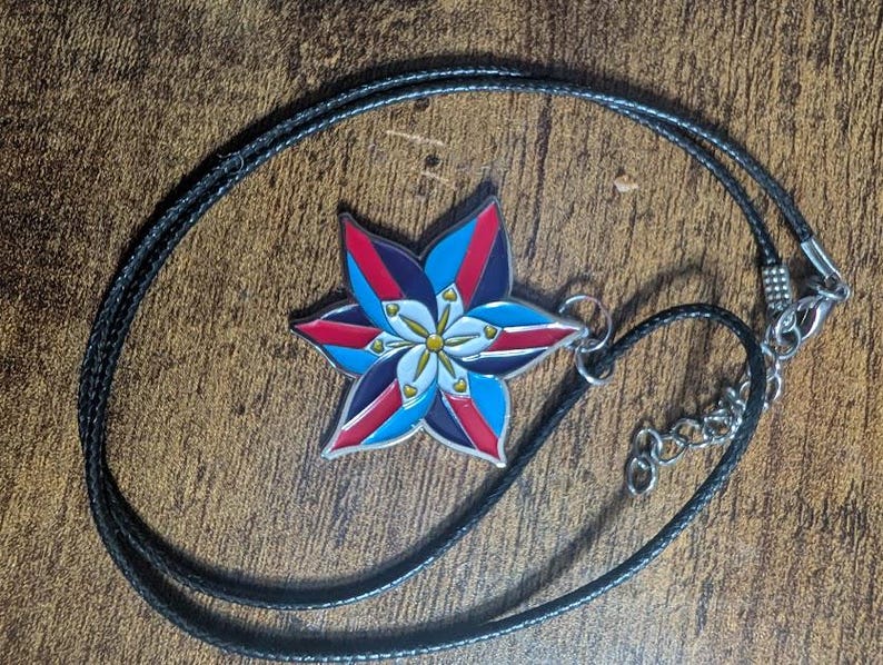 May include: A black cord necklace with a silver metal pendant featuring a colorful star-shaped flower design. The flower has red, blue, white, and yellow accents.