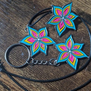 May include: Three flower-shaped pendants with a vibrant design. Each pendant features a teal outline, yellow petals, and pink centers. One pendant is attached to a keychain, and the others are on black cords. The pendants rest on a wooden surface.