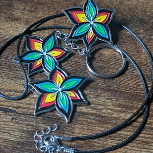 May include: A necklace and keychain featuring flower-shaped pendants. Each pendant has a silver outline and is filled with vibrant colors: red, yellow, green, and blue. The necklace has a black cord, and the keychain has a silver ring.
