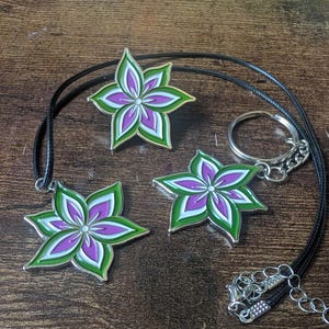 May include: A set of floral accessories, including a necklace, keychain, and pin. Each item features a flower design with purple, white, and green enamel. The necklace has a black cord, and the keychain has a silver ring.