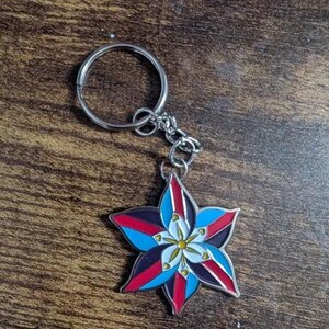 May include: A silver keychain with a blue, red, white, and black flower charm. The flower has a yellow center and is stylized to look like a star.