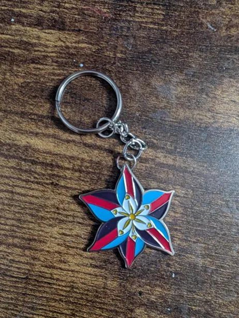 May include: A silver keychain with a blue, red, white, and black flower charm. The flower has a yellow center and is stylized to look like a star.