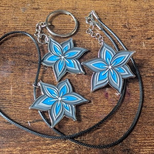 May include: Three flower-shaped pendants in gray, light blue, and white. One pendant is attached to a keychain, and two are on black cords. The pendants are on a wooden surface.