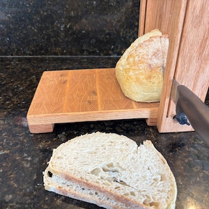 May include: A wooden bread slicer with a loaf of bread in place. A slice of bread is in the foreground. The slicer has a wooden base and a vertical wooden support with a stainless steel knife.