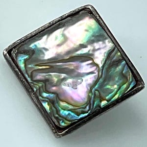 May include: Square silver-toned jewelry piece with a shimmering abalone shell inlay. The iridescent shell displays a mix of green, blue, purple, and silver hues. The shell is set within a square frame, creating a striking contrast.