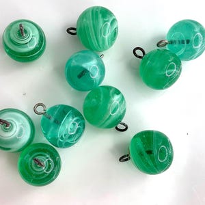 May include: A collection of ten round, green glass buttons with metal loops. The buttons have a swirled, marbled appearance with shades of light and dark green. The buttons are likely for sewing or crafting projects.