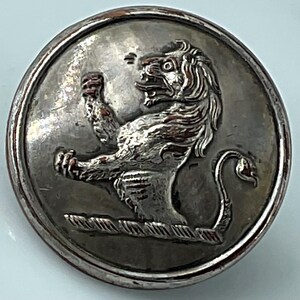 May include: A silver-toned button with a detailed relief of a lion rampant. The lion is depicted with a fierce expression, raised paw, and a curled tail. The button has a textured border and is likely made of metal.