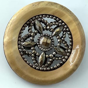 May include: A round button with a light brown outer ring and a silver-colored filigree center. The center features a starburst design with faceted, metallic-looking accents. The button has a vintage or antique aesthetic.