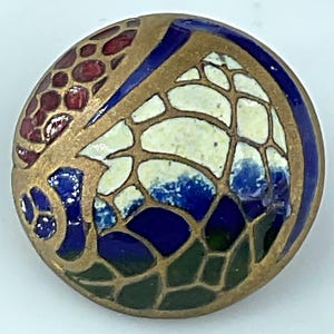 May include: A circular button featuring a cloisonné-style design with gold-toned metal outlines separating sections of enamel. The pattern includes segments of deep blue, off-white, dark green, and reddish-brown colors in a mosaic arrangement.