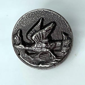 May include: A round, antique silver button with a detailed relief of a bird in flight over water. The button has a textured surface and a raised design, giving it a vintage appearance. The bird's wings are spread, and the button is likely made of metal.
