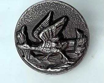 May include: A round, antique silver button with a detailed relief of a bird in flight over water. The button has a textured surface and a raised design, giving it a vintage appearance. The bird's wings are spread, and the button is likely made of metal.