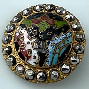 May include: A round, antique button with a gold-colored metal frame. The center features a colorful, abstract design in white, red, green, black, and blue. The button is encircled by a row of faceted, silver-colored stones.