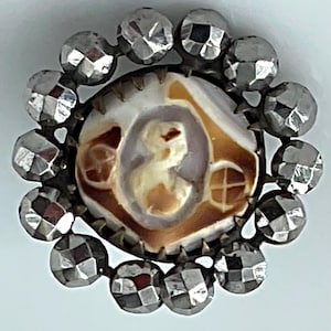 May include: A vintage brooch with a central cameo featuring a profile portrait in cream and brown tones. The cameo is surrounded by a ring of faceted, silver-toned stones, creating a circular design. The brooch has a detailed, antique aesthetic.