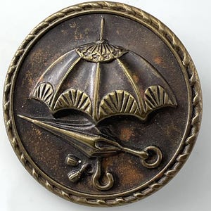 May include: A round, antique-style button with a raised umbrella design. The button has a bronze finish with a rope-like border. The umbrella is detailed with a curved handle and a decorative top.