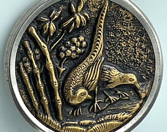 May include: A round, antique button with a detailed relief design. The button features two golden pheasants amidst bamboo stalks and foliage, set against a dark background. The button is framed by a silver-colored metal rim.