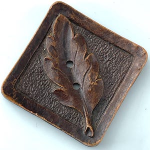 May include: A square-shaped button in a dark brown finish featuring a raised leaf design in the center. The button has a textured background within the square border and includes two vertical holes for attachment in the middle of the leaf.