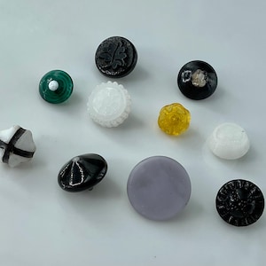 May include: A collection of various decorative buttons in different colors and designs. The buttons include a black button with a leaf design, a green button with a white center, and a white button with a scalloped edge.