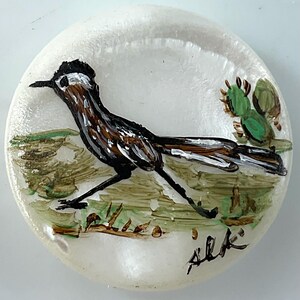 May include: A round, off-white, translucent stone disc featuring a hand-painted brown, black, and white roadrunner bird in profile. The bird stands on a green and brown painted landscape with a small green cactus and a black signature in the corner.