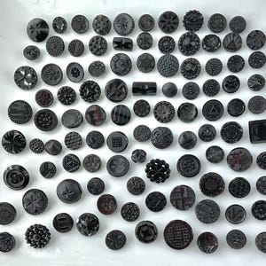 May include: A collection of various black buttons with different shapes and textures. The buttons include round, square, and geometric designs, with embossed patterns and star details. The buttons are arranged on a white surface.