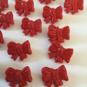 May include: A collection of small, glossy, red bow-shaped embellishments. These decorative bows are evenly spaced and appear to be made of a hard material. The bows have detailed folds and a classic bow tie design, perfect for crafting or embellishing projects.
