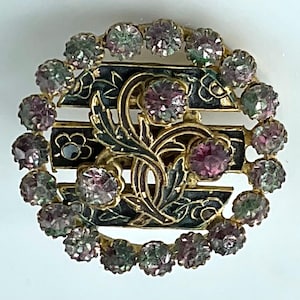 May include: A vintage-style brooch with a gold-tone metal frame. The brooch features a central design with black enamel and floral details. It is encircled by a ring of round, faceted, pink and green gemstones. A classic piece of jewelry.