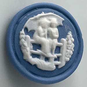 May include: A blue and white ceramic button with a raised design of two figures under an umbrella standing on a fence. The figures are in a white silhouette against a blue background.