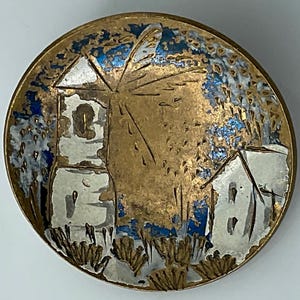 May include: A round, vintage button with a windmill and buildings design. The button features a gold-toned border with a central image of a white windmill and buildings against a blue and gold background. The button is likely made of metal.