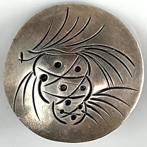 May include: A silver-toned, oval-shaped button or pendant with an etched design. The design features curved lines and circular shapes, creating an abstract floral or organic pattern. The metal has a slightly aged appearance.