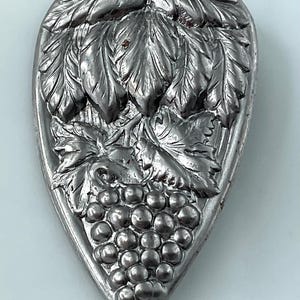 May include: A silver-colored, teardrop-shaped decorative item with a detailed grape and leaf design. The grapes are clustered at the bottom, with leaves and vines above. The item has a slightly aged appearance.