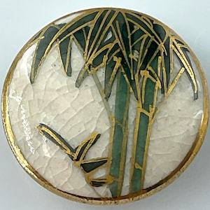 May include: A round, decorative button with a white, cracked background. The button features a green and gold bamboo design. The button has a gold-colored border.