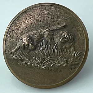 May include: A round, bronze-colored button with a detailed relief of two dogs in a grassy field. The dogs are depicted with textured fur and are surrounded by detailed foliage. The button has a textured background.