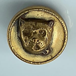 May include: A round, antique brass button with a detailed embossed image of a dog's face. The dog's face is centered within a circular frame, and the button has a rope-like border. The button has a vintage, worn appearance.