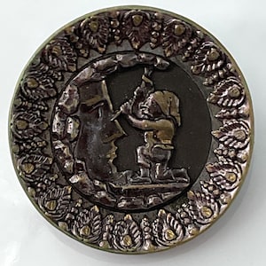 May include: A round, bronze-colored button with a raised design of a small figure chiseling a face into a rock. The button is surrounded by a decorative border of stylized leaves.
