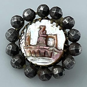 May include: A circular brooch featuring a central iridescent mother-of-pearl inlay depicting a silhouette of a figure standing near a structure. The center is surrounded by a border of twelve dark, faceted metallic beads arranged in a circular pattern.
