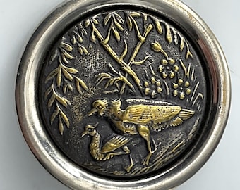 May include: A round, antique button with a silver rim. The button's center features a detailed relief of two birds, foliage, and flowers in gold against a dark background. The design is intricate and vintage-inspired.