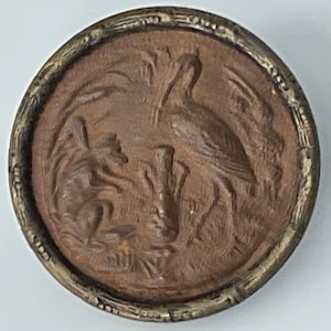 May include: A round, antique button with a detailed relief design. The central image depicts a bird and a small animal, possibly a rodent, amidst reeds or tall grass. The button has a brown center and a decorative, metallic border.