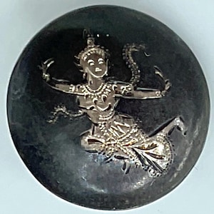 May include: A silver-colored pendant with a detailed relief of a dancing figure on a dark, circular background. The dancer is adorned with intricate jewelry and a headdress, with a snake-like element. The pendant has a polished finish.