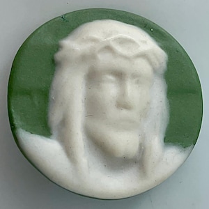 May include: A round, white cameo depicting a serene face with a crown of thorns, set against a green background. The cameo is a relief sculpture, showcasing detailed facial features and hair. The overall design is circular.