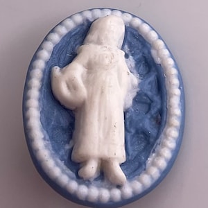 May include: An oval-shaped, blue and white cameo featuring a figure in a long white robe. The figure is set against a blue background with a floral design. The cameo is framed by a white beaded border.