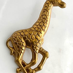 May include: A gold-colored metal giraffe brooch with a detailed textured pattern. The giraffe is depicted in a running pose with its head turned to the side.