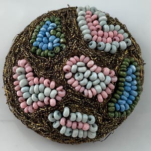 May include: A circular brooch featuring a textured gold metallic thread base adorned with clusters of small beads. The bead clusters are arranged in distinct patterns using light blue, pale pink, and olive green colors.