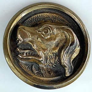 May include: A round, antique-style button with a detailed relief of a dog's head. The dog's head is gold-colored, set against a dark background, and framed by a brass-colored border. The button is likely made of metal.