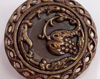May include: A round, ornate, brass button with a raised design of a grapevine with leaves and grapes. The button has a dark brown background and a silver-colored outer ring.