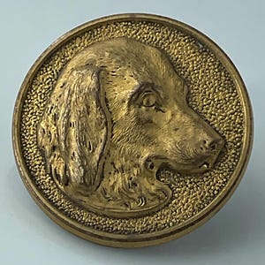 May include: A round, antique-style button with a detailed relief of a dog's head in profile. The dog is a golden color, set against a textured background within a circular frame. The button is likely made of metal.