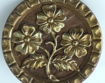 May include: Round, decorative button with a floral design. The button features three gold-colored flowers with detailed petals and leaves, set against a brown wooden background. The outer rim is a gold-colored, textured border.