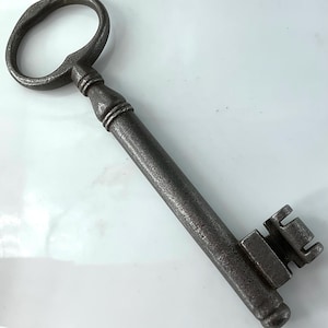 May include: An antique, oversized key with a dark, metallic finish. The key features a large, oval-shaped handle and a long, cylindrical shaft leading to a detailed bit. The key's design suggests a vintage or historical aesthetic.
