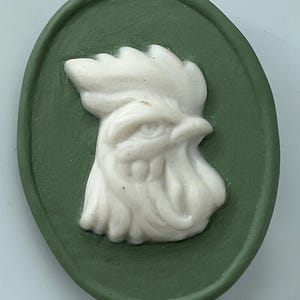 May include: An oval-shaped, green cameo featuring a white relief of a rooster's head. The rooster is detailed with a prominent comb and eye, set against a solid green background. The cameo has a smooth, polished finish.