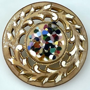 May include: A circular brooch featuring a gold-toned metal frame with an openwork leaf pattern accented by white enamel. The center is a round inlay filled with a mosaic of multicolored, irregular glass-like fragments in shades of blue, red, green, and yellow.