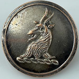 May include: A round, antique-style button with a detailed relief of a goat or ibex. The button has a tarnished silver finish with a raised bronze-colored goat figure in the center. The goat has detailed horns and fur.